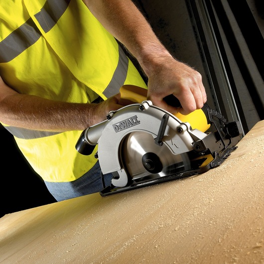 Person using a DEWALT circular saw to cut wood, wearing a yellow safety vest in a workshop setting.