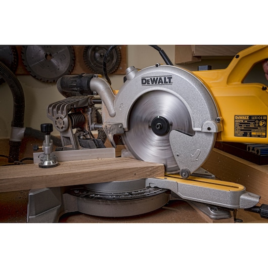 DEWALT mitre saw cutting wood in a workshop. The blade is spinning and sawdust is visible.