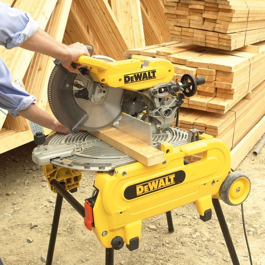 A person operating a DEWALT miter saw (model DT4210) to cut a piece of wood, with stacks of wooden planks in the background at a construction site.