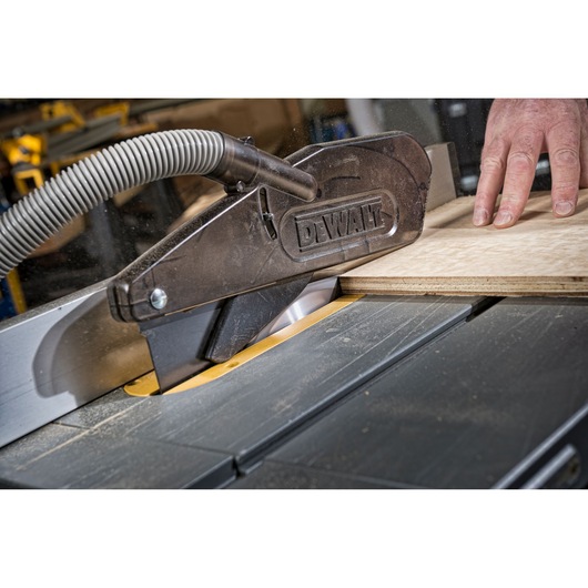 Close-up image of a DEWALT table saw, with a hand guiding a piece of wood through the blade guard. The saw is equipped with