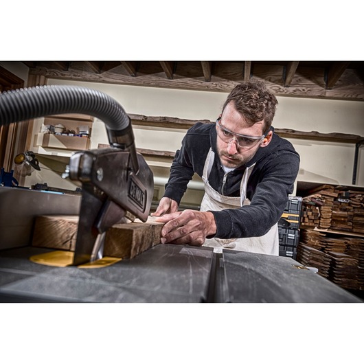 Person uses a DEWALT saw to cut wood in a workshop. Face is blurred. Wood stacks are visible in the background.
