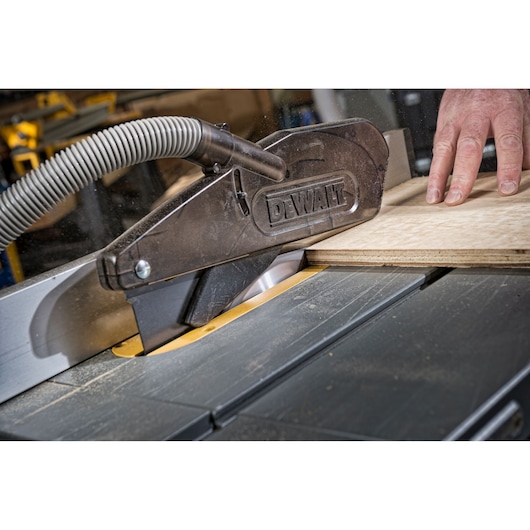 Close-up of a DEWALT table saw with a dust extraction hose, cutting a piece of plywood. A person's hand is guiding the wood