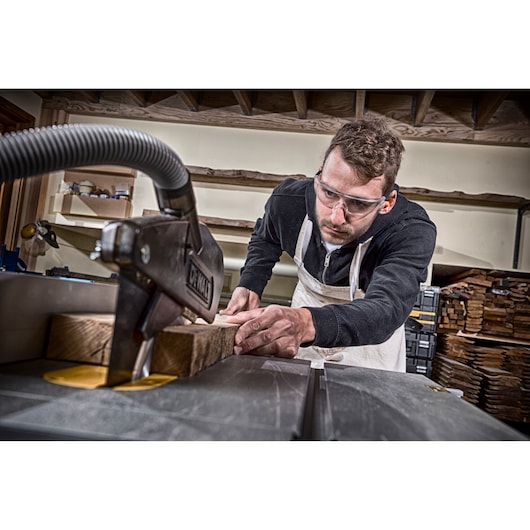 A person wearing an apron is using a DEWALT table saw to cut a piece of wood in a workshop. The person's face is blurred for