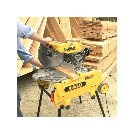 A person using a DEWALT miter saw to cut a piece of wood on a construction site, with stacks of lumber in the background.