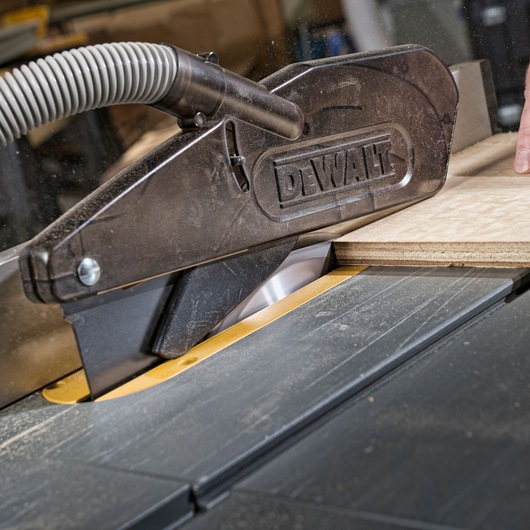 Close-up of a DEWALT table saw cutting a piece of plywood, showing the blade guard and dust extraction hose.