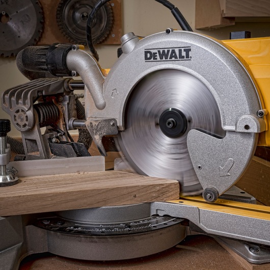 Close-up of a DEWALT miter saw cutting a piece of wood, with the circular blade in motion. The saw is placed in a workshop