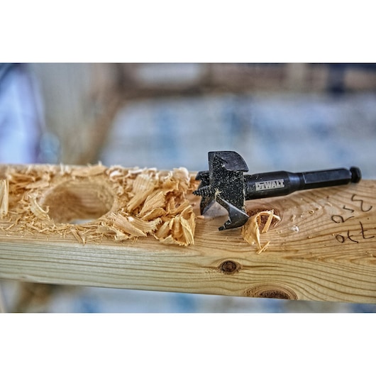 DEWALT wood drill bit with wood shavings and a drilled hole on a wooden plank in a workshop setting.