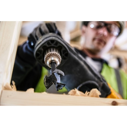 Close-up of a DEWALT power drill fitted with a wood drill bit in use, drilling through a wooden plank. The hand operating the drill is wearing a black glove. Background shows out-of-focus workshop setting.