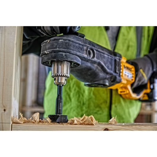 Close-up of a person using a DEWALT power drill with a 15mm bit to drill into a wooden board, creating wood shavings. The user is wearing gloves and a hi-vis vest.
