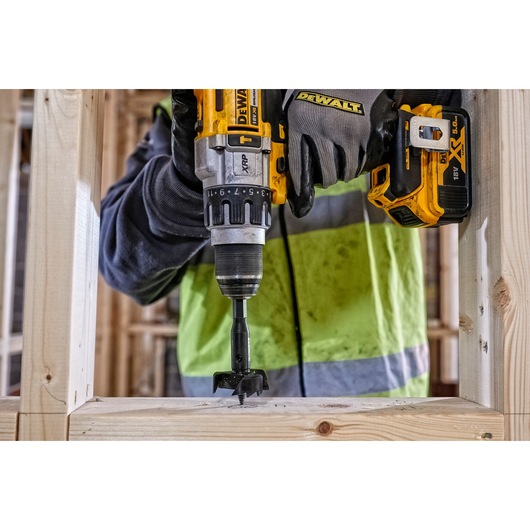 A person wearing work gloves and a high-visibility jacket is using a DEWALT cordless drill with a hole saw bit to drill into a wooden frame at a construction site.