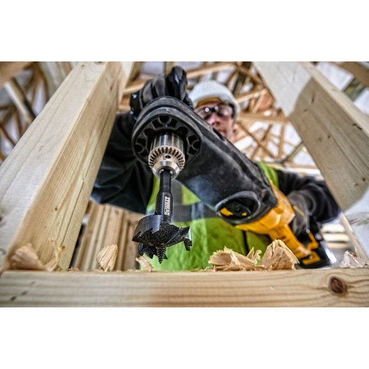 Close-up view of a DEWALT DT4585 drill bit in use, attached to a power drill, boring a hole through a wooden beam during construction. Wood shavings are visible around the area where the hole is being made.