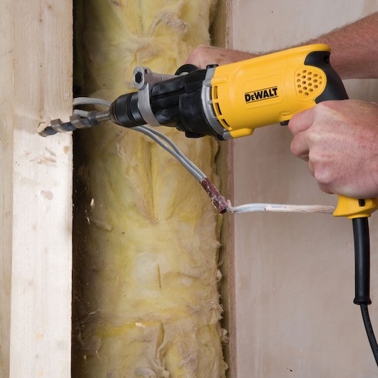 A DEWALT drill making a hole in wood near insulation, with wires visible in a construction setting.