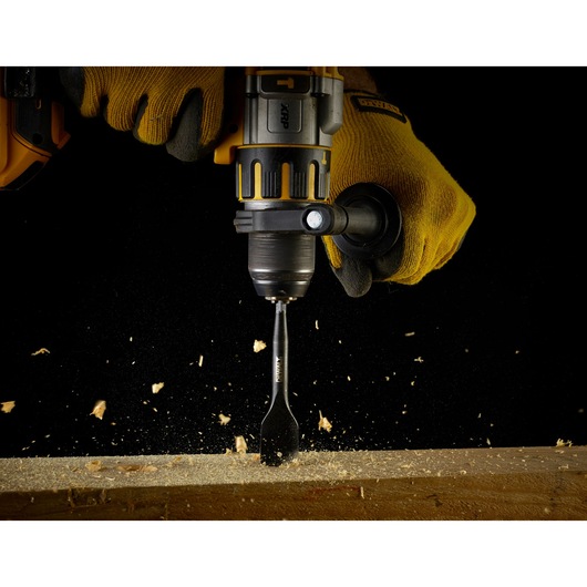 A close-up of a person wearing yellow gloves using a DEWALT electric drill with a flat wood drill bit to drill into a piece of wood. Wood shavings are flying as the drill operates.