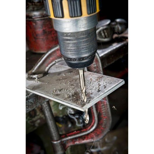 Close-up of a DEWALT drill with a metal drill bit, drilling through a metal plate held in place by a clamp.
