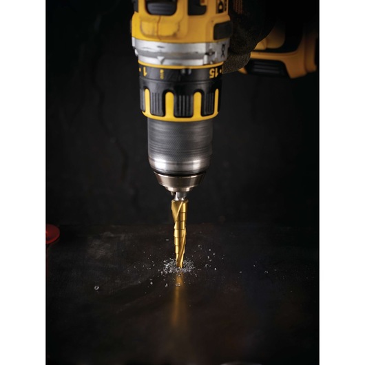 A close-up of a yellow and black DEWALT drill in use, drilling into a dark metal surface with a step drill bit. Metal shavings are visible around the drilling area.