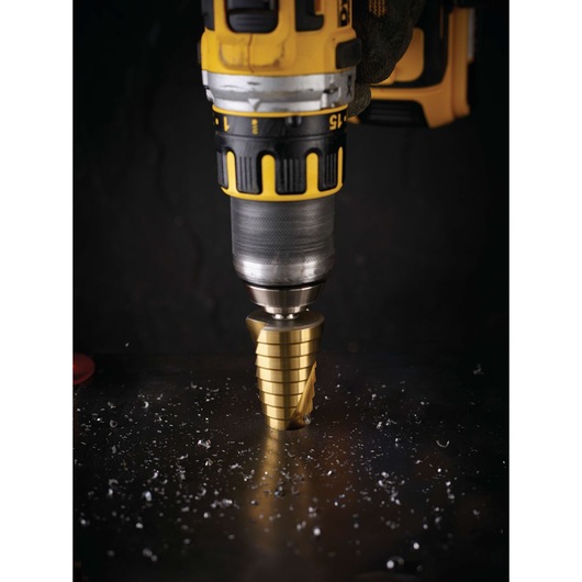 A close-up of a yellow and black cordless power drill using a gold step drill bit to drill into a metal surface, with metal shavings scattered around.