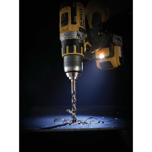 A gloved hand holding a DEWALT cordless power drill with a drill bit, actively drilling into a metal surface with metal shavings and debris visible beneath the bit, illuminated by a work light attached to the tool.