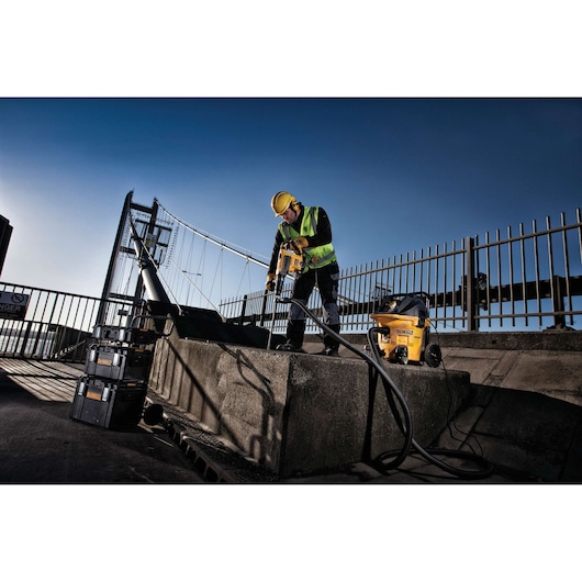 A construction worker wearing a yellow hard hat and high-visibility vest is using a DEWALT power tool to drill into concrete