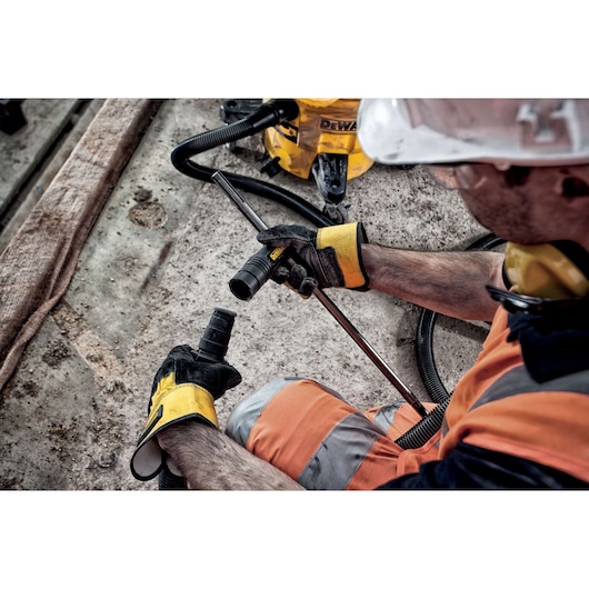 A worker wearing a hard hat, safety gloves, and an orange reflective vest assembles a DEWALT tool, possibly a drill bit or