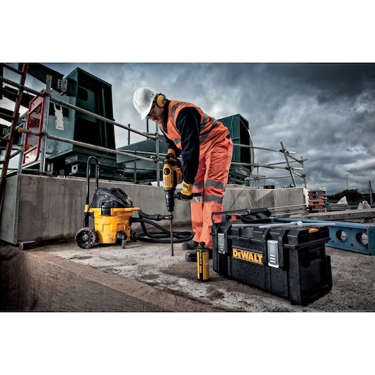 A construction worker wearing orange safety gear and a helmet uses a DEWALT power tool on a concrete surface. Nearby, there