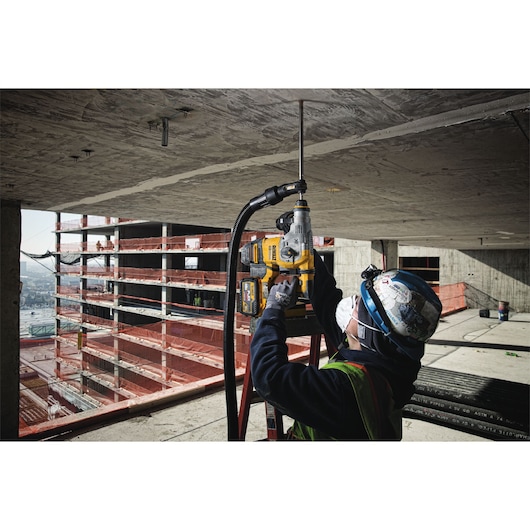 Construction worker using a DEWALT rotary hammer drill with a long drill bit to drill into a concrete ceiling at a building site. The worker is wearing safety gear including a hard hat, gloves, safety glasses, and a dust mask.