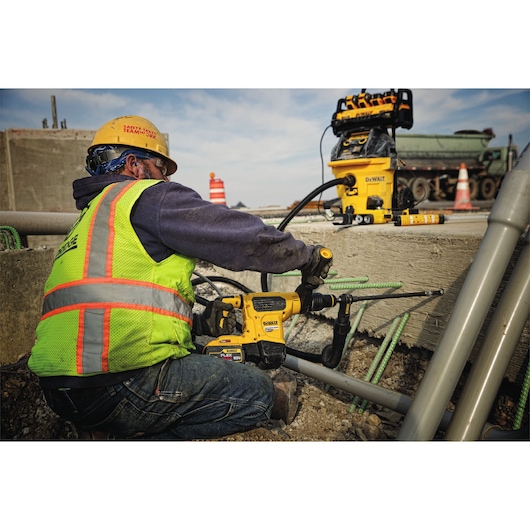 A construction worker wearing a safety vest and helmet uses a DEWALT rotary hammer drill to work on concrete at a construction site. Nearby, another DEWALT tool is placed on the concrete surface, with construction materials and safety cones visible.