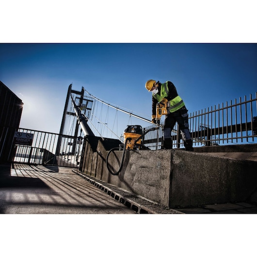 A construction worker wearing a yellow safety helmet and reflective vest is using a DEWALT power tool to drill into a