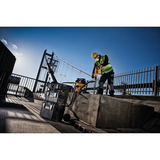 A construction worker wearing a yellow safety vest and hard hat uses a DEWALT power tool on a concrete surface outdoors