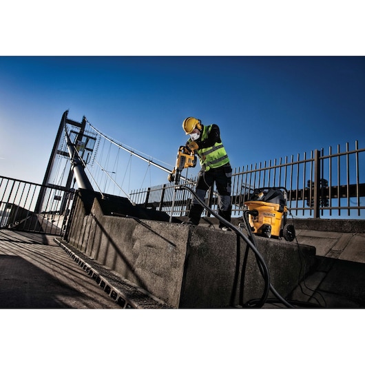 Person wearing safety gear using a DEWALT power tool and dust extractor on a concrete structure outdoors, with a suspension