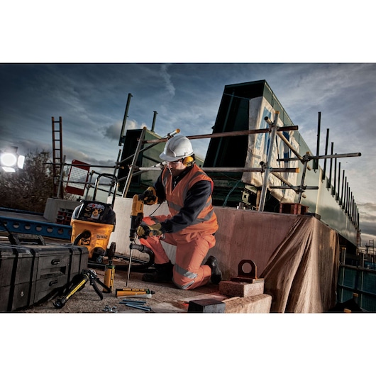 A construction worker wearing safety equipment, including a helmet and high-visibility vest, is using a DEWALT power tool