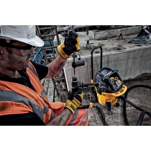 A worker wearing safety gloves and an orange reflective vest is operating a DEWALT power tool at a construction site. The