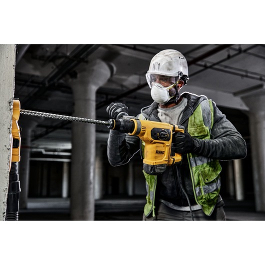 A worker wearing safety gear operates a yellow DEWALT rotary hammer drill to drill into concrete indoors.