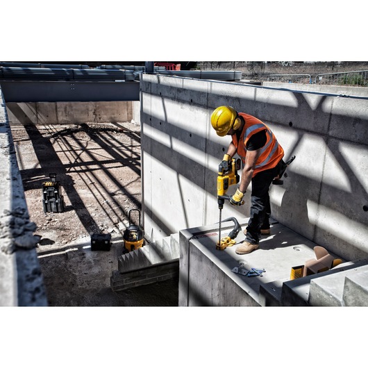 A construction worker wearing a yellow safety helmet and orange reflective vest is using a DEWALT power tool to drill into a concrete surface at a construction site.