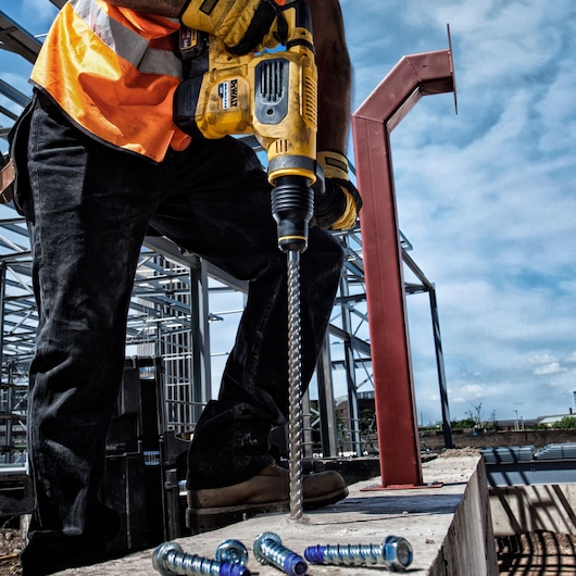 A construction worker wearing an orange safety vest and gloves uses a DEWALT rotary hammer drill (DT60814) to drill into concrete on a building site, with metal bolts visible in the foreground.