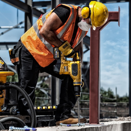 A construction worker wearing a hard hat, ear protection, safety glasses, face mask, gloves, and an orange reflective vest, is operating a yellow DEWALT power drill to bore into concrete at a construction site.
