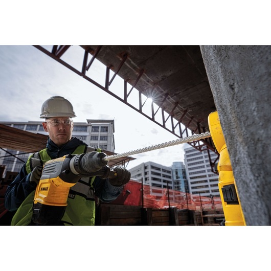 A person wearing a safety helmet and vest uses a DEWALT power drill with a long drill bit on a construction site. Modern buildings and orange construction barriers are visible in the background.