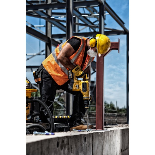 A construction worker wearing a yellow hard hat, ear protection, and an orange safety vest is using a yellow DEWALT power drill to drill into a concrete surface at an industrial site with steel framework in the background.