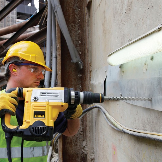 A construction worker wearing a yellow hard hat, safety gloves, and ear protection is using a DEWALT DT60816 rotary hammer drill to drill into a concrete wall. The worker is operating the tool close to a metal fixture and fluorescent light, with various pipes and wires visible in the background.