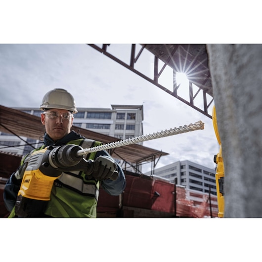 Worker uses DEWALT DT60817 drill bit at construction site, wearing safety gear. Urban buildings in background.