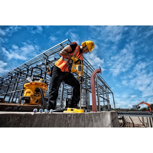 A construction worker wearing a yellow hard hat and orange safety vest operates a DEWALT power drill at a construction site, with steel framework and a clear blue sky in the background. DEWALT equipment and tools are visible nearby.
