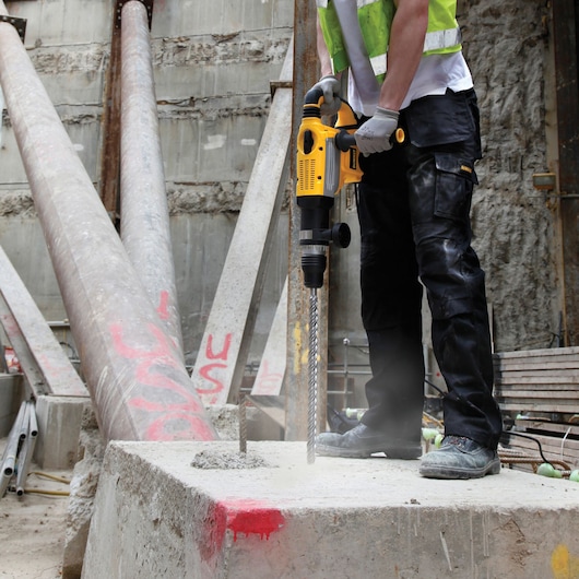 A construction worker using a yellow DEWALT rotary hammer drill to drill into a concrete block at a construction site. The worker is wearing safety gloves, a reflective vest, and protective work pants.