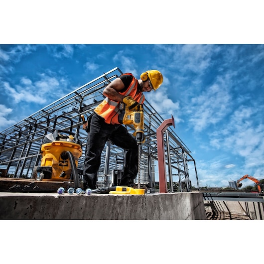 A construction worker wearing a yellow hard hat and orange safety vest uses a DEWALT power tool to drill into concrete at an outdoor construction site. Steel framework of a building and construction equipment are visible in the background.