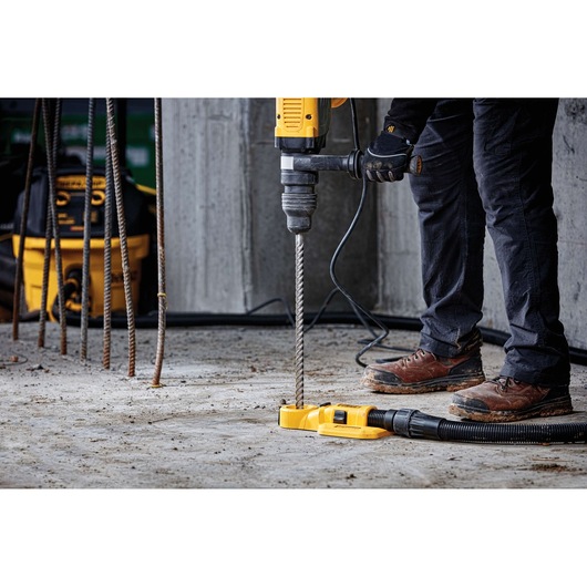 A person wearing work boots and gloves is using a yellow DEWALT hammer drill with a long drill bit to drill into a concrete floor at a construction site. Several pieces of rebar and a DEWALT tool bag are visible in the background.