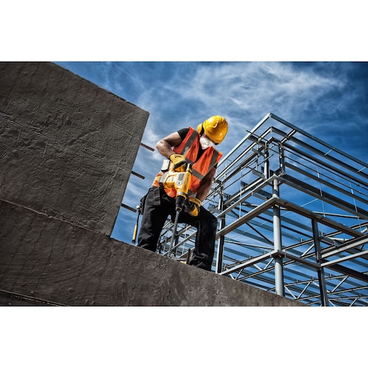 A construction worker wearing a yellow hard hat, orange safety vest, and face mask is using a yellow DEWALT power tool to drill into a concrete surface at an outdoor construction site with a metal framework structure in the background.