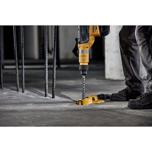 A person using a yellow DEWALT rotary hammer drill with a spiral bit to drill into a concrete floor at a construction site. Several vertical steel rebars are visible in the background.