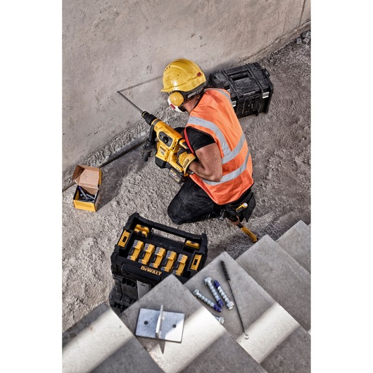 A construction worker wearing a yellow hard hat, orange reflective vest, and ear protection is using a yellow DEWALT power drill to drill into a concrete wall. The worker is kneeling on the ground next to a DEWALT tool box, another black tool case, and an open box of drill bits. Construction hardware and accessories are visible on nearby concrete steps.