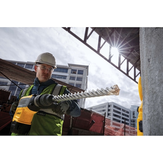 A construction worker wearing a hard hat and safety vest operates a DEWALT rotary hammer drill with a long, spiral drill bit at an outdoor urban construction site. Modern buildings and sunlight are visible in the background.