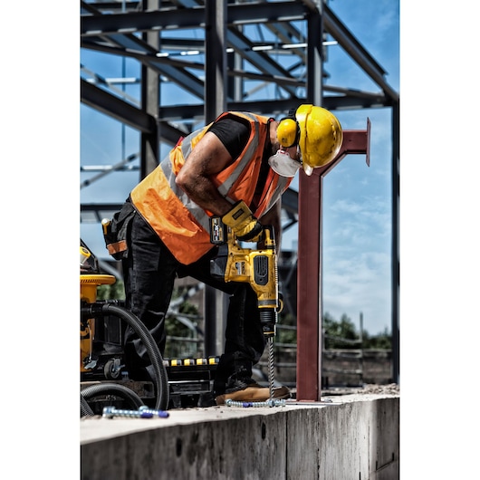 Construction worker wearing an orange safety vest, yellow hard hat, ear protectors, and a face mask is using a DEWALT power drill to drill into concrete at a building site. Steel framework of the building is visible in the background.