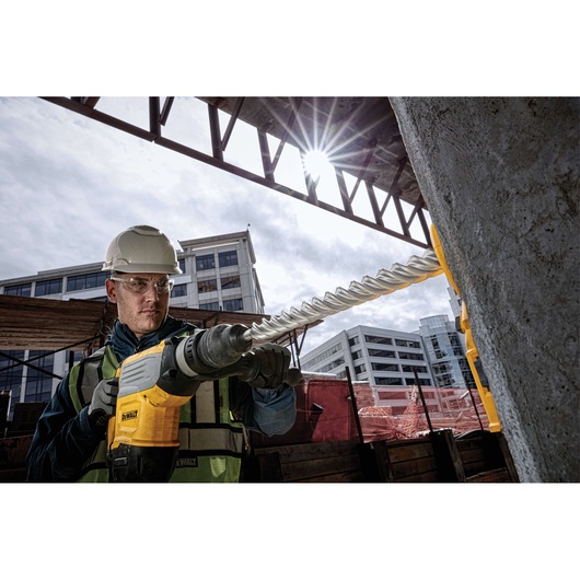 A construction worker wearing a hard hat and safety vest operates a DEWALT power drill with a long spiral bit to drill into concrete at an outdoor construction site, with buildings visible in the background.