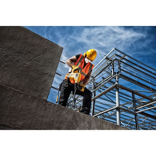 A construction worker wearing a yellow hard hat, safety vest, and face mask uses a DEWALT power tool to drill into a concrete structure at a building site with steel framework under a blue sky.